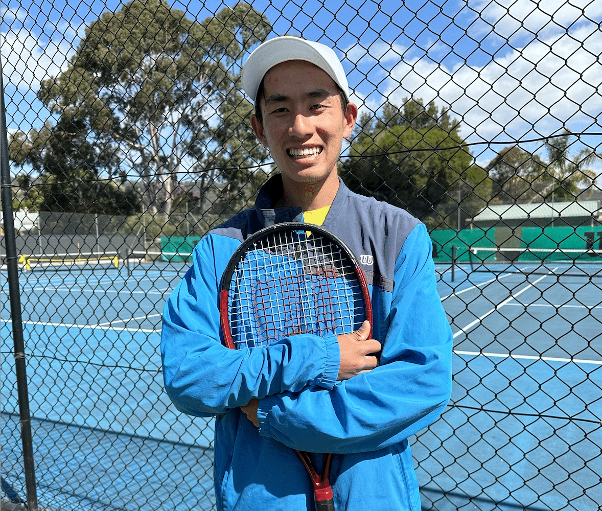 Aaron is dressed at Wilson clothing at Denman Tennis Club. He is smiling and hugging his racquet.