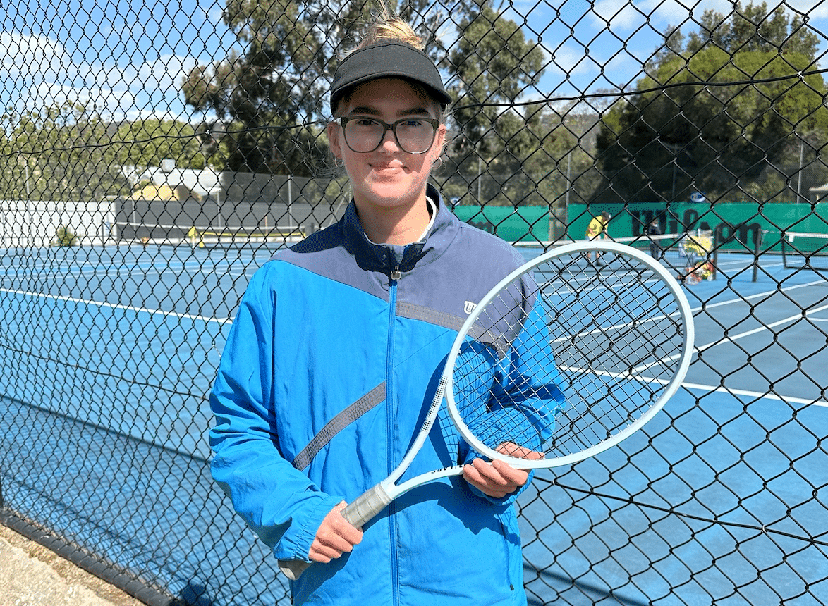 Sienna is holding her racquet across her body and smiling at the camera. She is dressed in WIlson clothing at Denman Tennis Club
