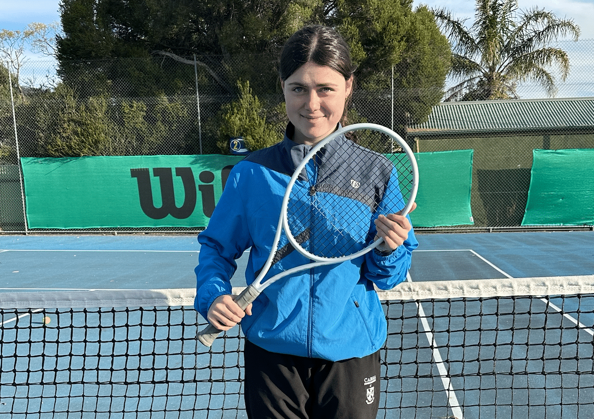 Ash is wearing a blue Wilson jacket on court at Denman Tennis Club. She is holding her racquet across her body and smiling